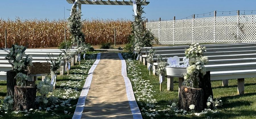 Outdoor ceremony aisle under a flower arch with white chairs and a cornfield beyond