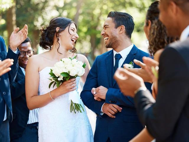 Smiling bride holding white bouquet beside her groom in a navy suit as guests applaud outdoors
