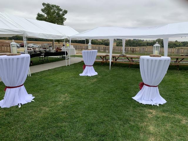 Tall white-draped cocktail tables with red sashes set on lawn beneath a white event tent