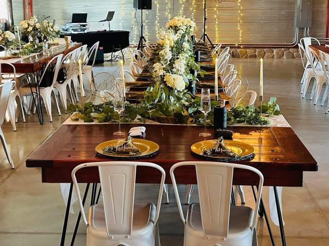 Long wooden farm table decorated with white florals and greenery, surrounded by white metal chairs under string lights