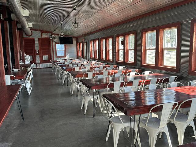 Rows of long wooden tables and white chairs set in a bright barn hall with tall windows