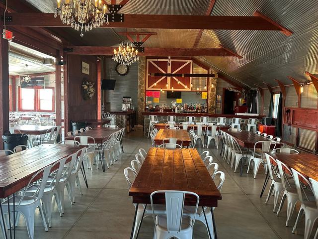 Red barn interior with a long wood table, white chairs and chandeliers hanging from the beams