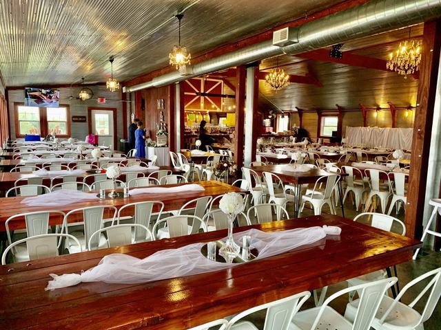 Large rustic reception hall with long wooden tables, white chairs, chandeliers, and a bar