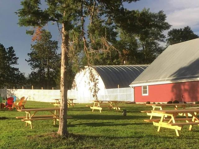 Green lawn with yellow picnic tables, tall trees and a red barn beside a white building