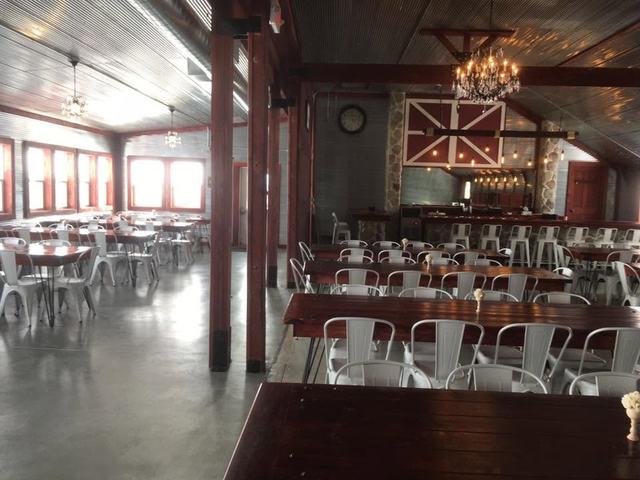 Large barn hall with tall windows, long wooden tables, white chairs, and a chandelier overhead