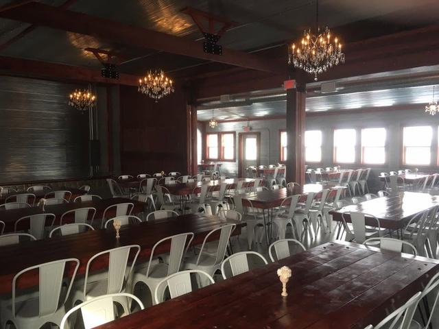 Open hall with chandeliers hanging over farm tables and white chairs near tall windows