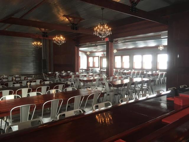 Wide reception hall with chandeliers over long farm tables, white chairs, and bright windows