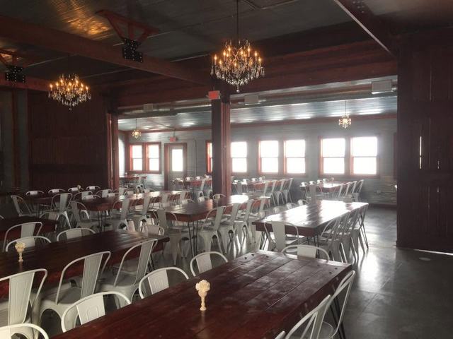 Event hall with exposed wood beams, chandeliers, long farm tables, and white chairs